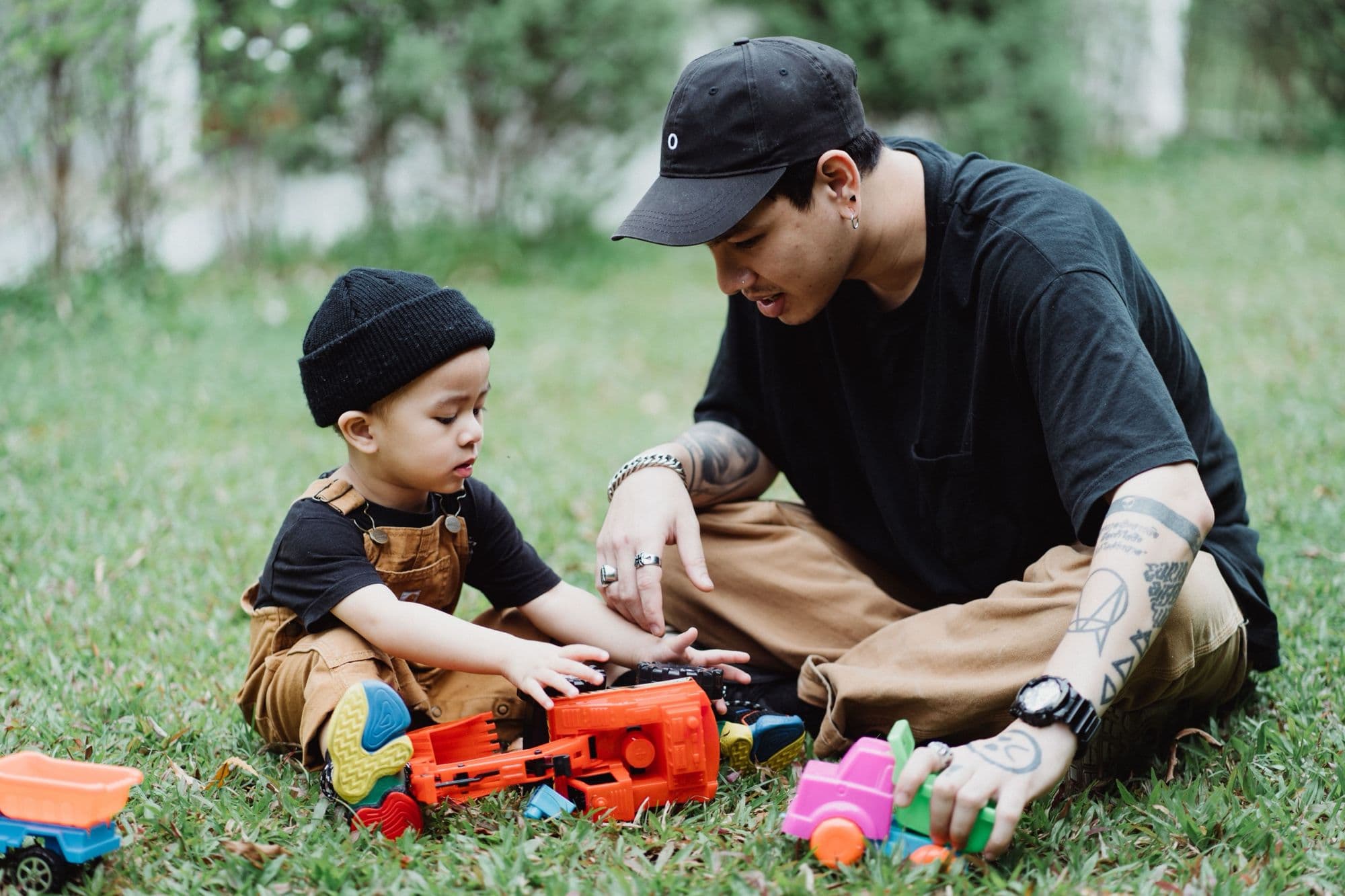 A father in a cap sits cross-legged on the grass, playing with toy trucks alongside his young son.
