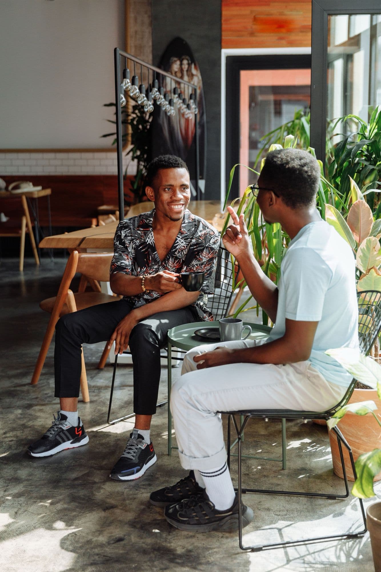 Two men sit across a small table in a sunlit café, coffee in hand, one smiling as the other talks.